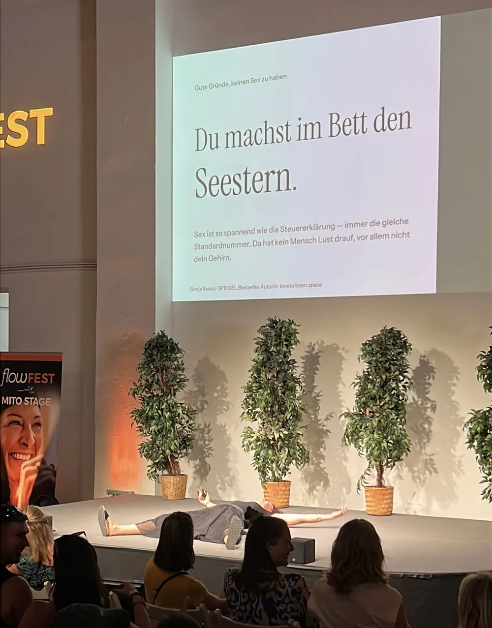 Der Seestern auf der Bühne des Flowfest: Sonja Ruess liegt ausgestreckt auf der Bühne, im Hintergrund die Folie „Du machst im Bett den Seestern.“